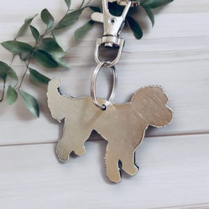 May include: Silver metal keychain shaped like a doodle dog. The keychain has a silver ring and a silver clip.