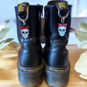 May include: Pair of black leather boots with yellow stitching and platform soles. Each boot features a skull charm with a red bow attached to the pull loop. The charms are silver with a white skull design.