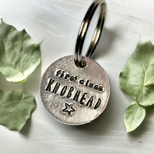 May include: Silver keyring with a round tag that says "first class knobhead" with a star below the text.