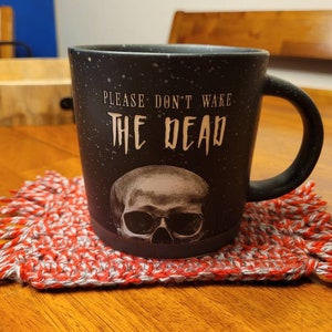 May include: Black coffee mug with a skull graphic and the text "PLEASE DON'T WAKE THE DEAD". The mug is sitting on a red and white crocheted coaster.