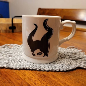 May include: White ceramic mug with a black silhouette of Maleficent from Disney's Sleeping Beauty. The mug is sitting on a gray crocheted coaster.