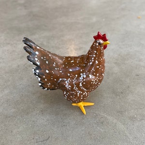 May include: A brown and white speckled chicken figurine with a red comb and yellow feet. The chicken is standing on a gray surface. This decorative item is a farm animal figurine.