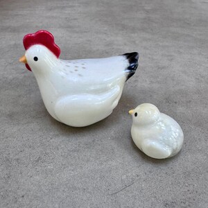 May include: Two ceramic figurines: a white hen with a red comb and black tail feathers, and a small white chick. The hen has a yellow beak and the chick has a small black eye. The figurines are likely decorative.