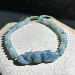 May include: A jade bracelet featuring a carved lotus flower charm. The bracelet is made from light green jade beads and has a green string fastening.