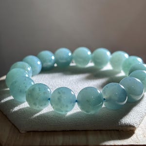May include: A bracelet composed of large, spherical, pale blue beads threaded onto a blue elastic cord. The beads have a translucent quality and a speckled pattern. The bracelet is presented on a light grey surface.