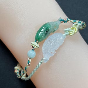 以下が含まれることがあります： 緑と透明の2つの魚の形をしたチャームが付いた翡翠のブレスレット。ブレスレットは、緑、金、茶色の色合いの編み込みコードで、小さな青いビーズが付いています。魚のチャームは精巧に彫刻されています。