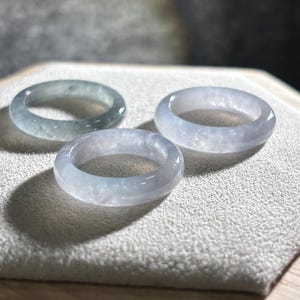 Könnte beinhalten: Drei transluzente Jade-Ringe in Blau- und Grüntönen werden auf einer strukturierten Oberfläche präsentiert. Die Ringe haben eine glatte, polierte Oberfläche, die die natürlichen Farb- und Texturvariationen des Jades hervorhebt.