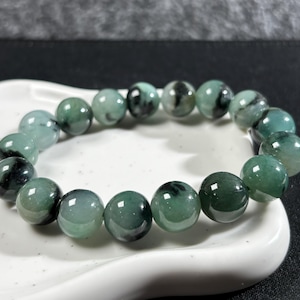 May include: A bracelet with polished, round beads in green and black hues. The bracelet is presented on a white, curved surface. The beads are approximately 1.3 cm in diameter, strung together to form a complete circle, suitable for wrist wear.