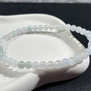 Puede incluir: Una delicada pulsera compuesta por pequeñas cuentas redondas blancas y verde pálido. Las cuentas están ensartadas para formar una banda flexible. La pulsera se muestra sobre una superficie blanca y curvada.