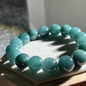 May include: A stretch bracelet composed of smooth, round, polished beads in a vibrant teal-green color. The beads exhibit a glossy, reflective surface with subtle variations in tone, arranged in a circular formation to form a continuous loop.