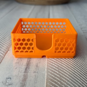 May include: Bright orange, rectangular desk organizer with a honeycomb pattern on the sides. The organizer has a slot on one side and is made of a solid, possibly 3D-printed material. The design is modern and functional.