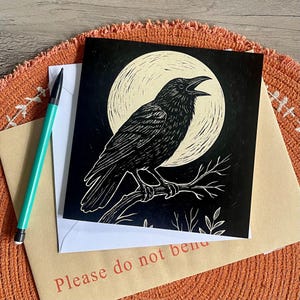 May include: A black and white art print of a raven perched on a branch in front of a full moon. The print is on a square card, resting on an envelope with the text "Please do not bend". A teal pen is also visible.