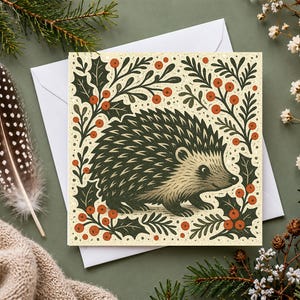 May include: A square greeting card featuring a dark green hedgehog illustration surrounded by holly leaves and red berries on a cream background, resting on a white envelope.