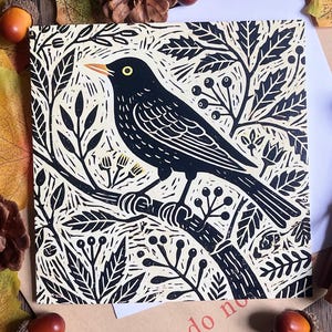 May include: A square print featuring a black bird with a yellow eye and orange beak perched on a branch. The design uses a linocut style with black ink on a cream background, depicting surrounding foliage, leaves, and small berries in a repeating pattern.