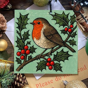 May include: A square greeting card featuring a linocut-style illustration of a robin perched on a holly branch. The bird has a bright orange breast and brown wings, set against a light green background with dark green holly leaves and red berries.