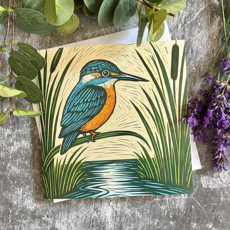 May include: A greeting card featuring a detailed illustration of a kingfisher bird perched on a branch. The bird has teal and orange plumage, set against a backdrop of reeds and water. The artwork is in a woodcut style.