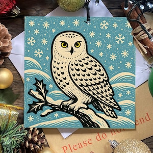 May include: A square print featuring a cream-colored snowy owl with black markings perched on a dark branch against a light blue background with white snowflakes and stylized snow drifts, rendered in a bold, graphic block-print style.