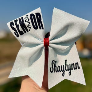 May include: A white glitter hair bow featuring a red center band. The left side displays the black text SENIOR 2024, and the right side features the black script name Shaylynn. The bow has a structured, multi-layered design with pointed ends.