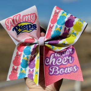 May include: A pink and white cheer bow with glitter. One side features a pink, yellow, blue, and purple pattern of Peeps with the text "All my Peeps". The other side features the text "I wear cheer bows" in purple.