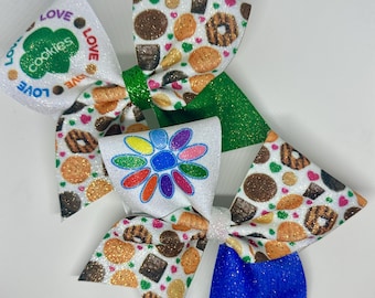 May include: Three decorative hair bows. One bow features the text "Love Cookies" and a green emblem. The other two bows have a pattern of cookies and hearts. The bows have glitter accents in green, white, and blue.