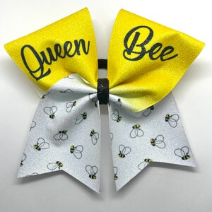 May include: A yellow and white cheer bow with glitter. The bow has a white side with a pattern of black and yellow bees and a yellow side with the words "Queen Bee" in black.