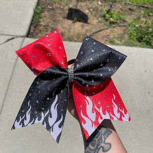 May include: A black and red cheer bow with white flames and silver glitter. The bow is decorated with rhinestones.