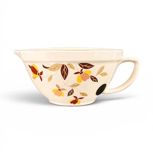 May include: A cream-colored ceramic mixing bowl with a pouring spout and a side handle. The exterior features a repeating floral pattern in shades of brown, yellow, and soft pink, with thin brown stems and leaves connecting the flower motifs.