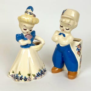 May include: Two vintage ceramic figurines. One is a woman in a blue dress and hat, holding a basket. The other is a man in a blue uniform with a hat, serving as a vase. Both have closed eyes and floral accents.