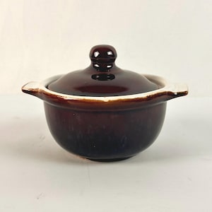 May include: A small, round, dark brown ceramic bowl with two side handles and a matching domed lid. The bowl features a glossy, deep brown glaze that transitions to a lighter, creamy white tone along the upper rim and the handle edges.
