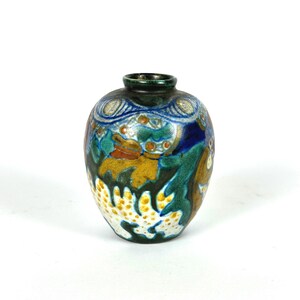 May include: A small, decorative ceramic vase with a round body and a dark green rim. The vase features a colorful design with blue, green, yellow, and white patterns. The design includes stylized floral or abstract motifs.