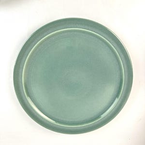 May include: A round, light teal-colored ceramic plate. The plate has a slightly raised rim and a smooth, matte finish. The plate is designed for serving food.