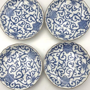 May include: A set of four round ceramic plates featuring a white base with a blue floral pattern of swirling vines and blossoms. Each plate has a scalloped edge and a thin metallic rim, displaying a consistent decorative design across all pieces.