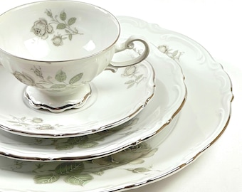 May include: A set of white porcelain teacups and saucers with a delicate floral design featuring gray roses and green leaves. The edges of the cups and saucers are trimmed with a silver band.