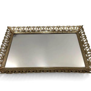 May include: A rectangular vanity tray featuring a mirrored base and an ornate gold-tone metal frame. The raised border displays a repeating openwork scroll pattern with intricate detailing along the edges, providing a reflective surface.
