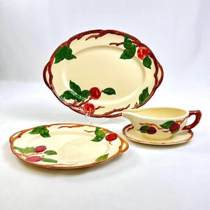 May include: A set of three vintage ceramic serving dishes featuring a cream-colored background with a red apple and green leaf design. The set includes a large oval platter, a smaller oval platter, and a gravy boat with a matching saucer.