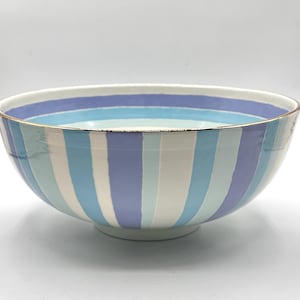 May include: A ceramic bowl featuring vertical stripes in shades of lavender, light blue, and white. The bowl has a smooth, rounded shape with a gold-toned rim along the top edge, providing a clean and simple design for serving various food items.
