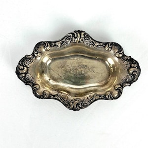 May include: An oval silver-toned metal tray featuring a scalloped edge with ornate embossed floral and scrollwork detailing. The center of the tray has a smooth, reflective surface with an engraved monogram script visible in the middle.