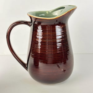 May include: A brown ceramic pitcher with a wide, curved handle. The pitcher has a dark brown exterior with horizontal lines and a light green interior. The rim is a lighter brown color. The pitcher is designed for serving beverages.