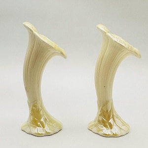 May include: Two cream-colored ceramic vases shaped like calla lilies. The vases have a curved, trumpet-like form with textured vertical lines and a flared opening. The base of each vase has a leaf-like design with a golden hue.