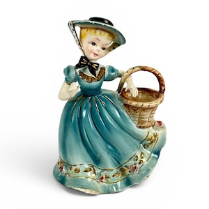 May include: A vintage ceramic figurine of a woman in a teal dress and hat, holding a woven basket. The dress has gold floral accents. The woman has blonde hair and a black bow. The figurine is on a white background.
