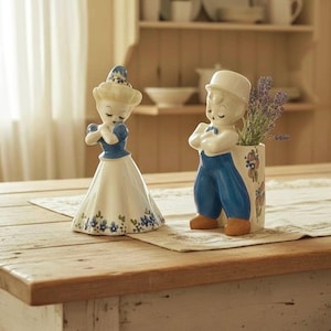 May include: Two ceramic figurines, a woman and a man, in traditional Dutch attire. The woman wears a blue and white dress, while the man has blue pants and a white top. A vase with lavender is next to the man.
