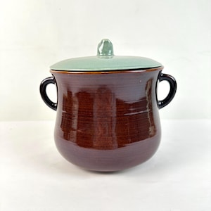 Puede incluir: Una olla de cerámica marrón con tapa. La olla tiene dos asas negras y una tapa verde claro con un pequeño pomo. El exterior de la olla tiene un acabado brillante.