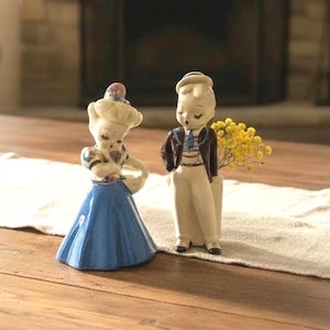 May include: Two ceramic figurines, a woman in a blue dress and a man in a suit, stand side-by-side. The woman wears a white hat and holds a napkin. The man has a small vase of yellow flowers next to him. The figurines are on a wooden surface.