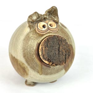 May include: A ceramic pig-shaped bottle stopper with a cork nose. The pig is a light brown color with darker brown accents and has two large, round eyes. The stopper is designed to fit into a bottle opening.