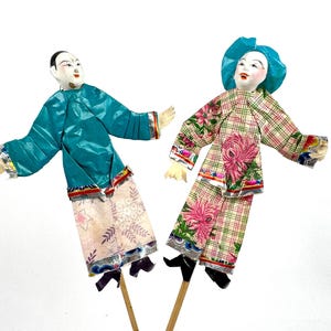 May include: Two vintage Chinese paper puppets on sticks. The puppet on the left is wearing a teal blue jacket and pink pants with floral designs. The puppet on the right is wearing a plaid jacket with pink flowers and pink pants with a plaid pattern. Both puppets have painted faces.