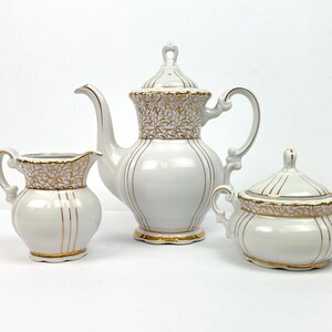 May include: A white porcelain tea set with gold accents. The set includes a teapot, creamer, and sugar bowl. The teapot has a tall, curved spout and a decorative lid. The creamer and sugar bowl have matching gold trim. The set is on a white background.