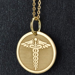 May include: A gold necklace with a round pendant featuring a medical symbol, the caduceus, with a snake wrapped around a staff with wings.