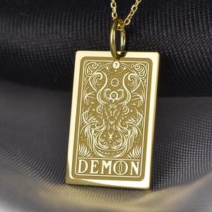 May include: A gold-colored pendant necklace featuring a rectangular charm with an intricate engraved design and the word "DEMON" at the bottom. The pendant hangs from a delicate gold chain. The background is a dark, textured fabric.