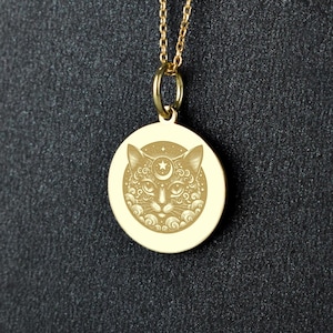 May include: Gold-colored pendant necklace featuring a detailed cat face design within a circular frame. The pendant hangs from a delicate chain with small, round beads. The cat's face includes a star and crescent moon. The necklace is displayed against a dark background.