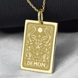 May include: Gold-colored pendant necklace with a rectangular pendant. The pendant features an engraved design of a horned creature with the word "DEMON" at the bottom. The necklace is displayed on a black and white background.